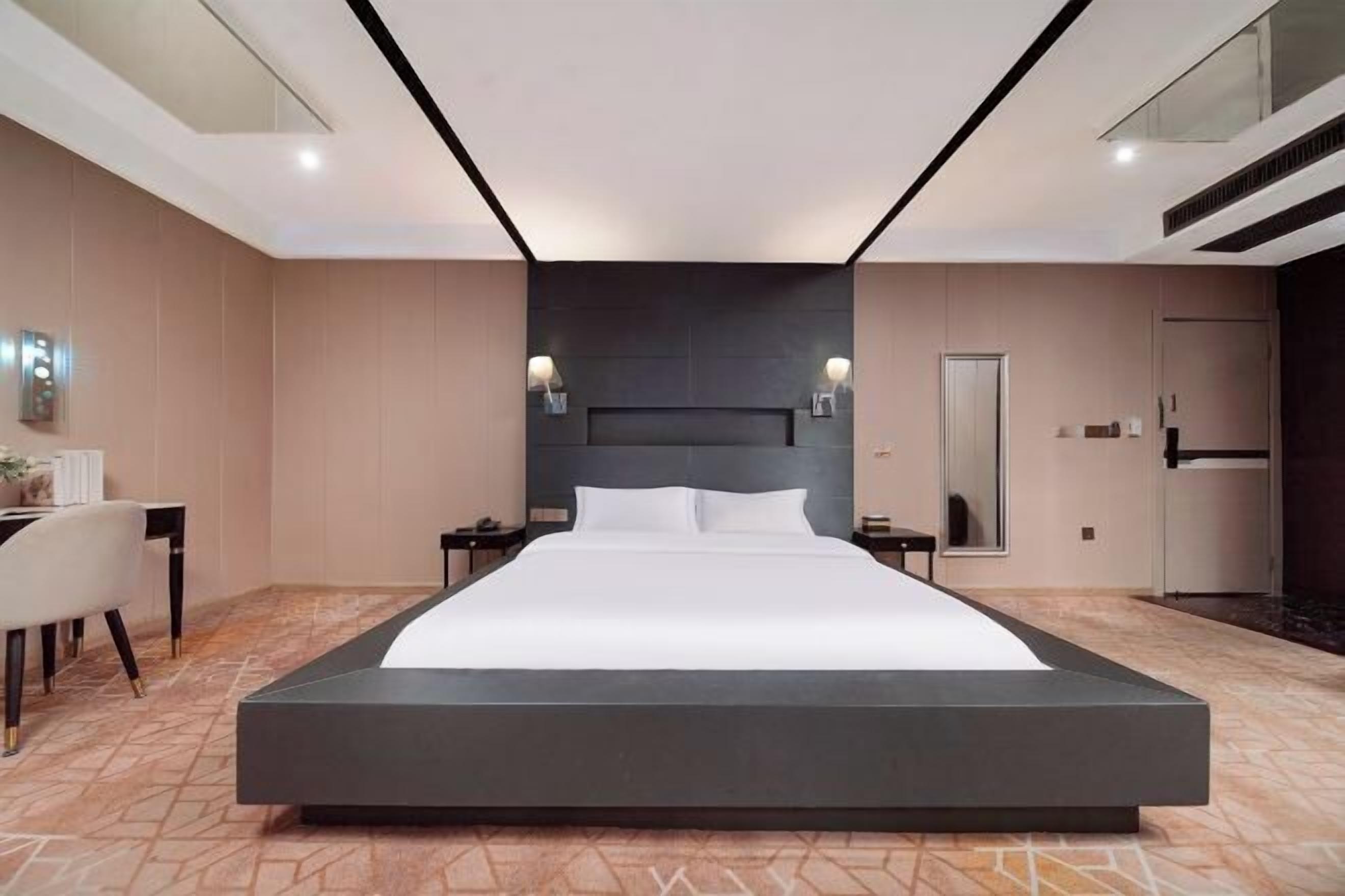 A modern hotel room with a large bed, a desk with a chair, and a wardrobe.