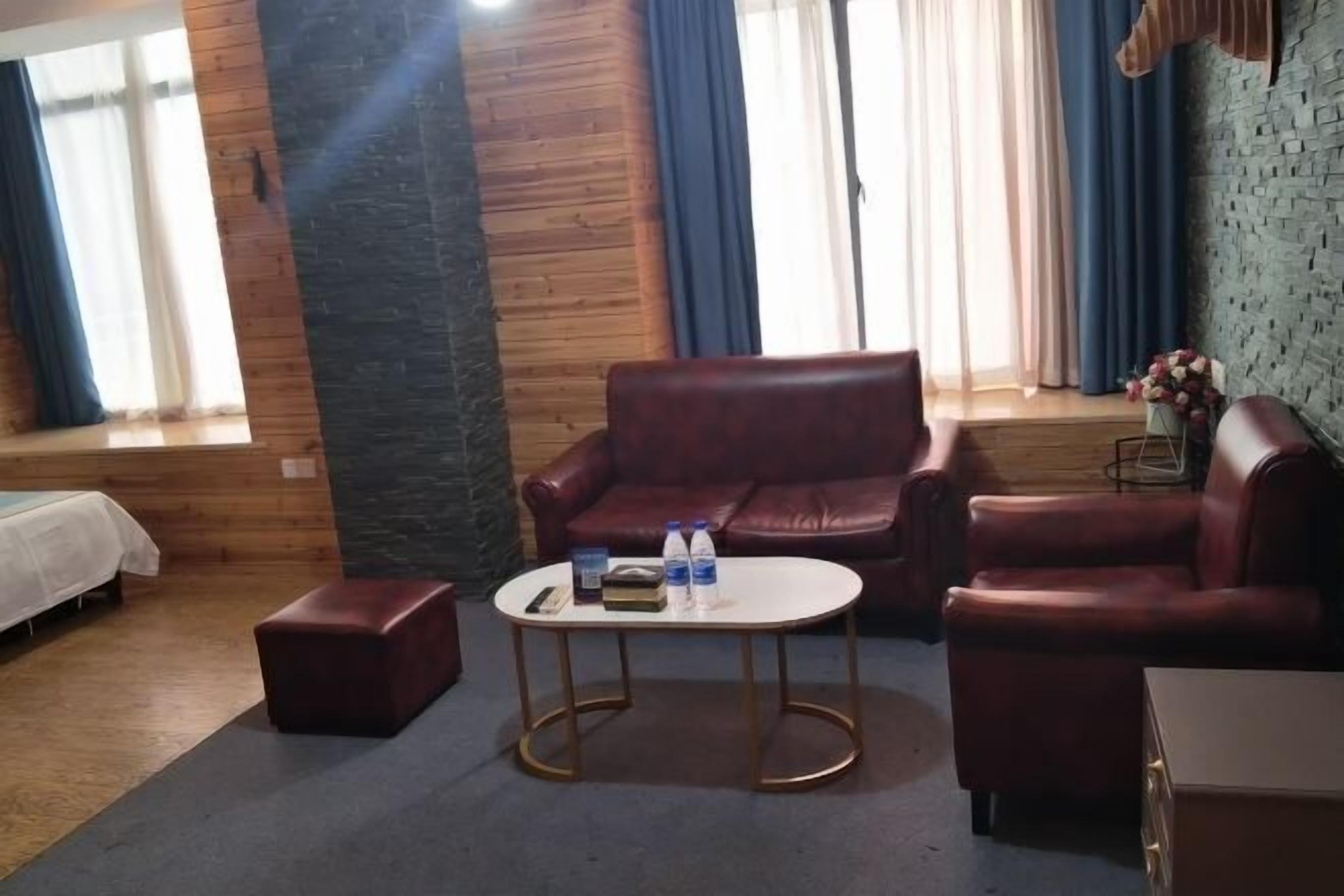 A cozy room with a wooden wall, a red leather sofa, a small round table with a book and bottles, and a window with curtains.