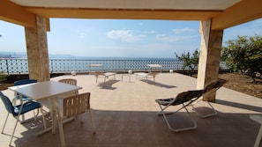 Een overdekt terras met uitzicht op zee, met een tafel, stoelen en een lounge fauteuil.