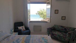 Un dormitorio con cama, una silla, un sofá, una ventana con vista al mar y un aire acondicionado de pared.
