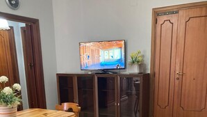 Un salon comprenant une table à manger en bois, des chaises, un canapé avec des coussins verts et turquoise, un téléviseur posé sur un meuble et une porte en bois.