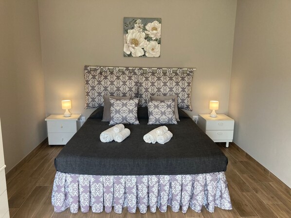 A neatly made bed with a dark bedspread and matching curtains, flanked by two bedside tables with lamps, and a floral painting above the bed.