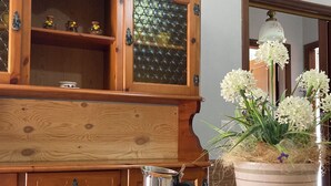 Un buffet en bois avec des portes en verre, une cafetière en métal, des tasses et des soucoupes blanches, et un pot rayé orné de fleurs blanches.