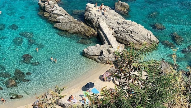 Une zone côtière caractérisée par une eau turquoise et limpide, des formations rocheuses et une plage de sable dotée de parasols.