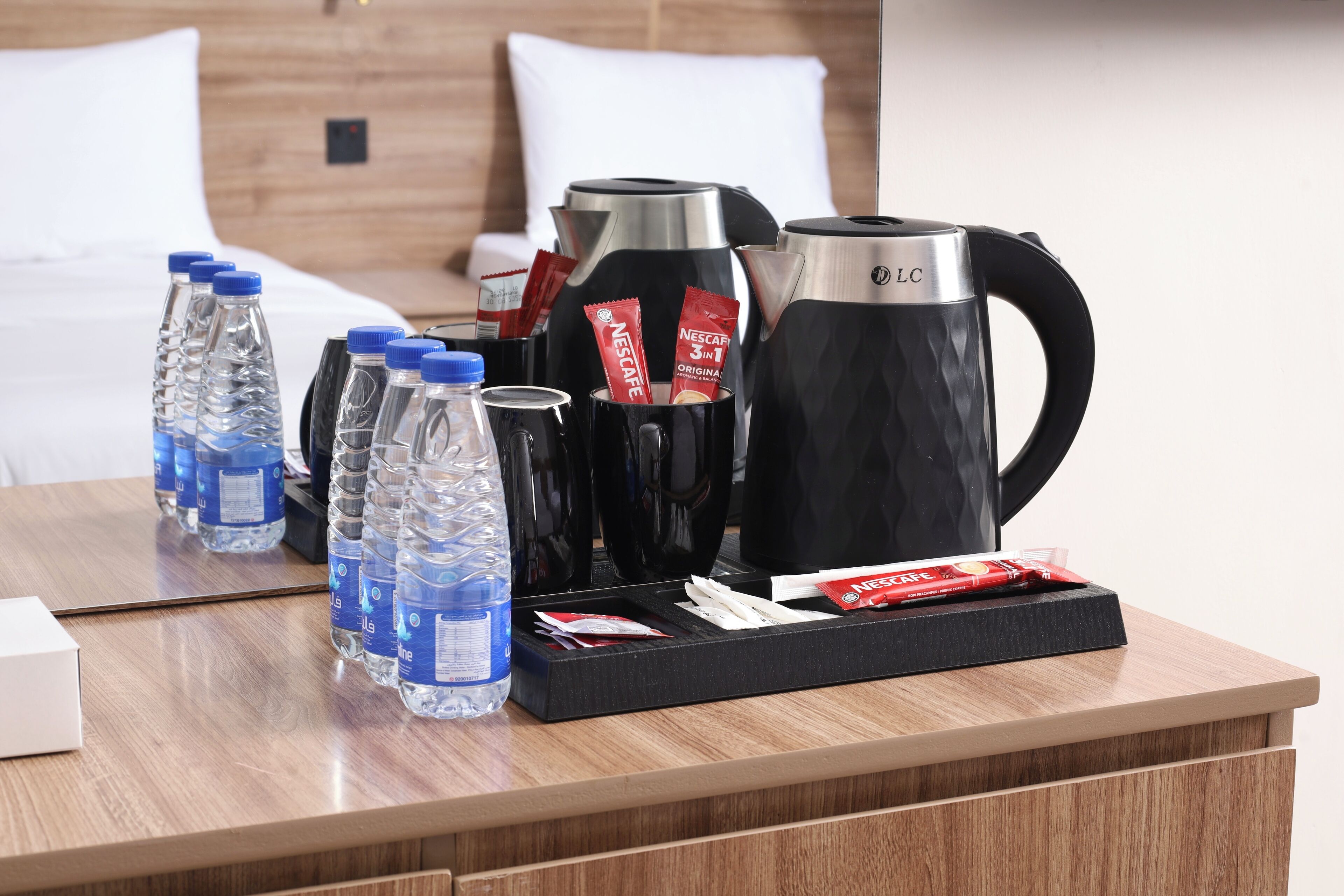 Hotelkamer met houten hoofdeinde, een bed met witte lakens, een houten bureau met waterflesjes, een koffiezetapparaat en een dienblad met koffiepads.