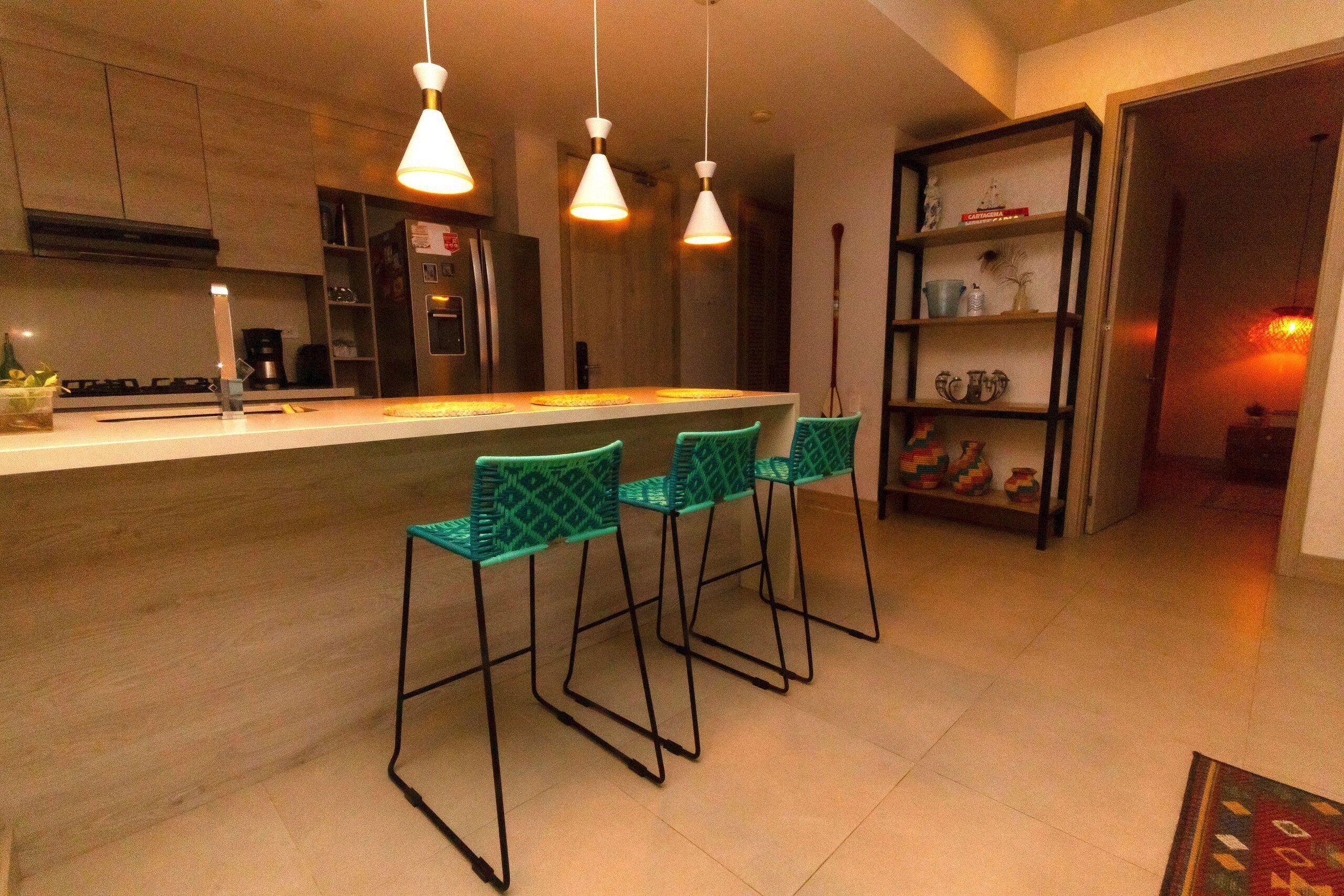 A modern kitchen with a central island, bar stools, pendant lighting, and a built-in bookshelf.