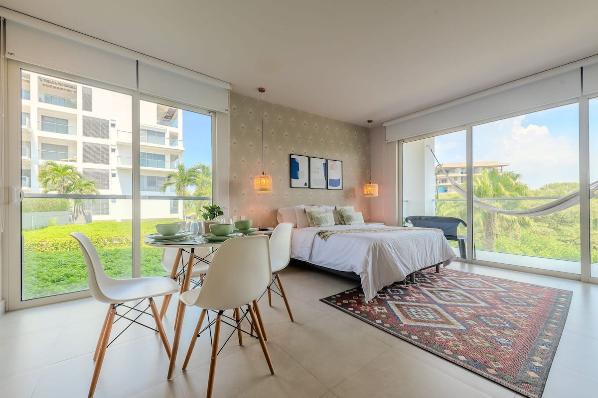 A modern bedroom with a large bed, a dining area with chairs, and a view of a building and greenery outside.