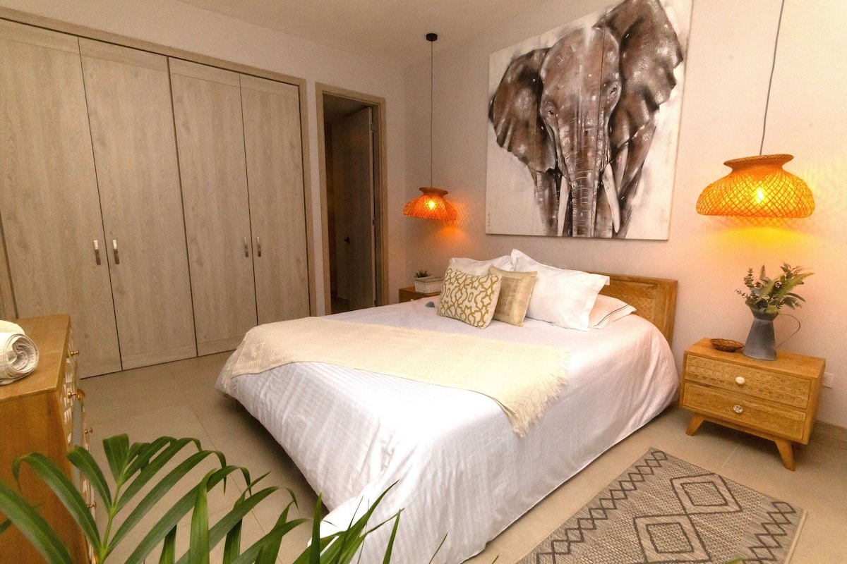A bedroom with a large bed, a wardrobe, a bedside table with a lamp, and a wall-mounted artwork of an elephant.