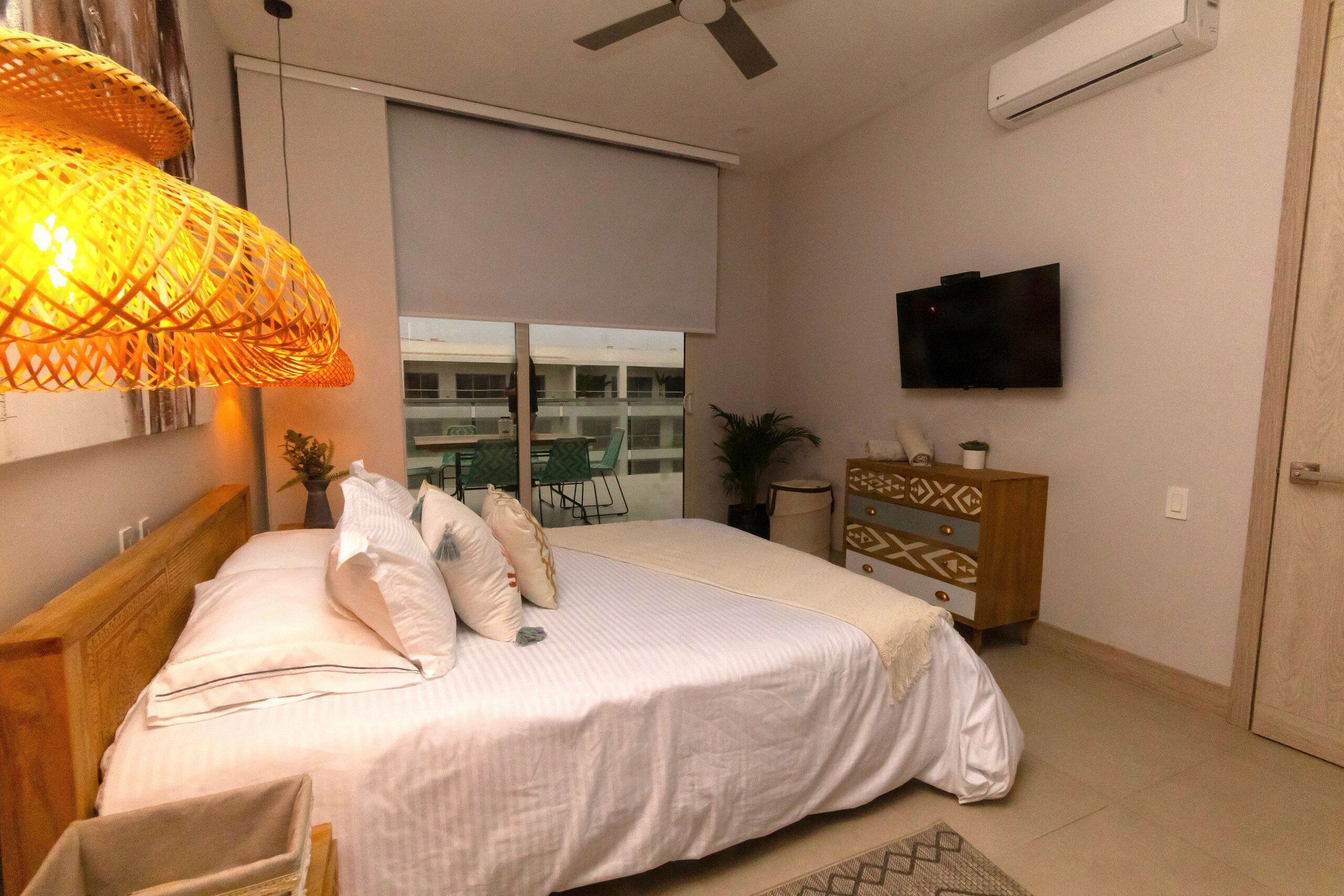 Een slaapkamer met een bed, een televisie, een plafondventilator en een raam met uitzicht op gebouwen.