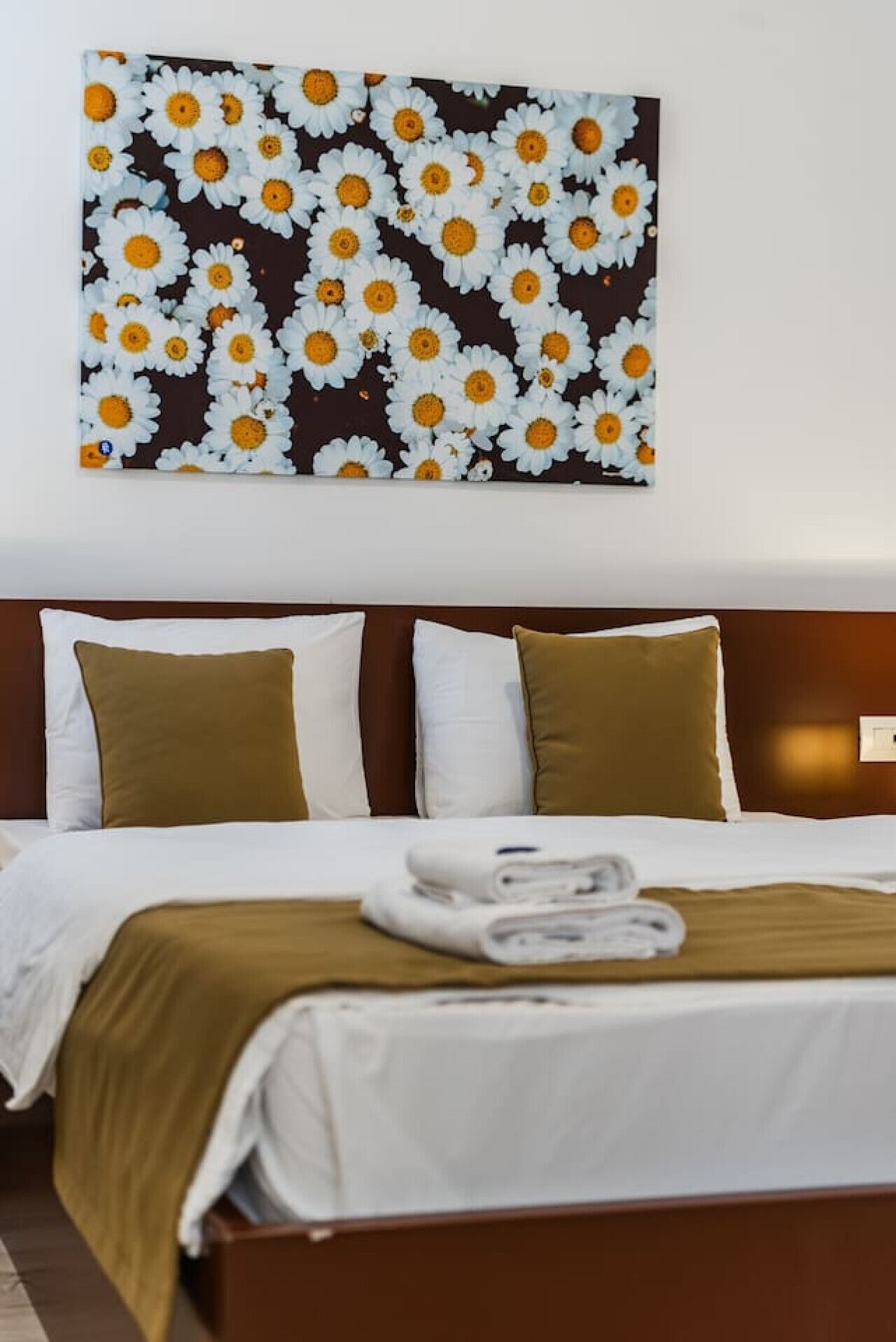 Une chambre d’hôtel avec un lit, deux oreillers, une couverture et une œuvre d’art représentant des marguerites, accrochée au mur.