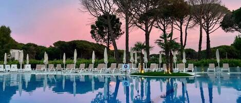 Ein Pool mit Liegestühlen und Sonnenschirmen, umgeben von Bäumen und blauem Himmel.