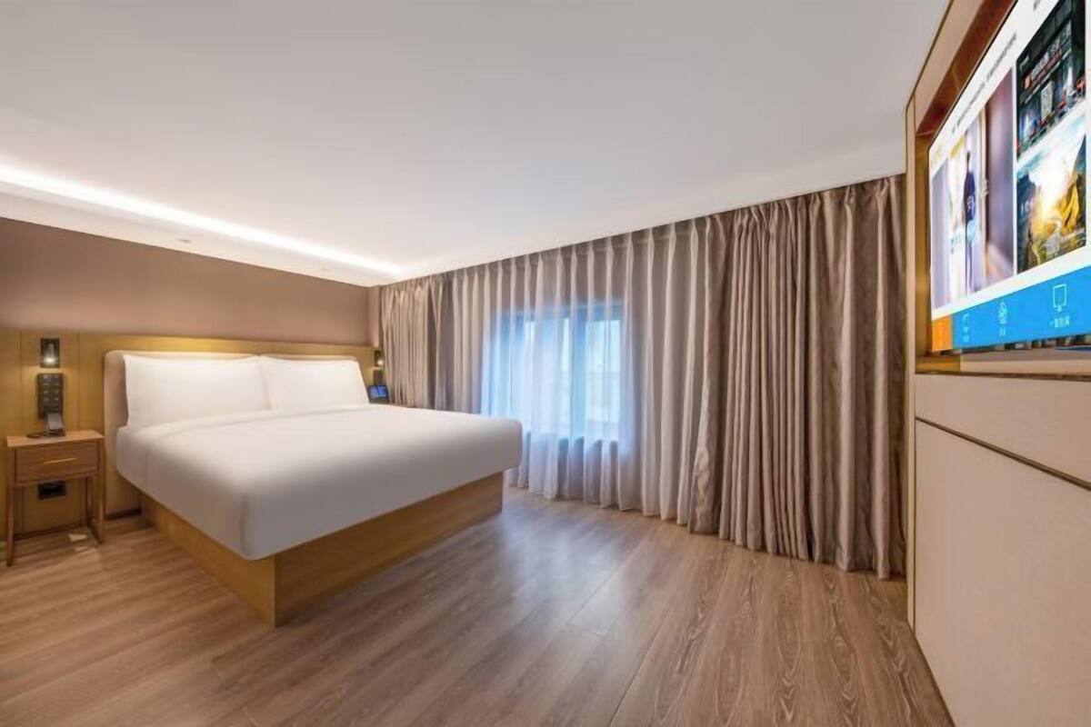 Quarto de hotel com uma cama grande, piso de madeira, mesa de cabeceira, televisão e janela com cortinas.