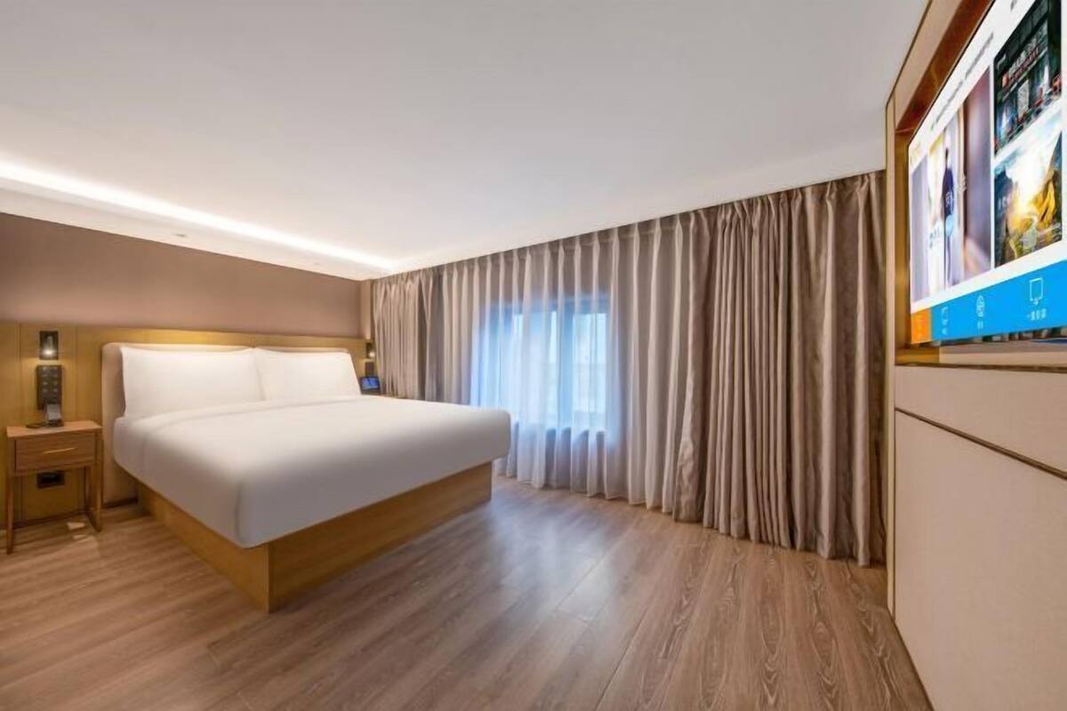 Quarto de hotel com cama, piso de madeira, mesa de cabeceira, televisão e janela com cortinas.