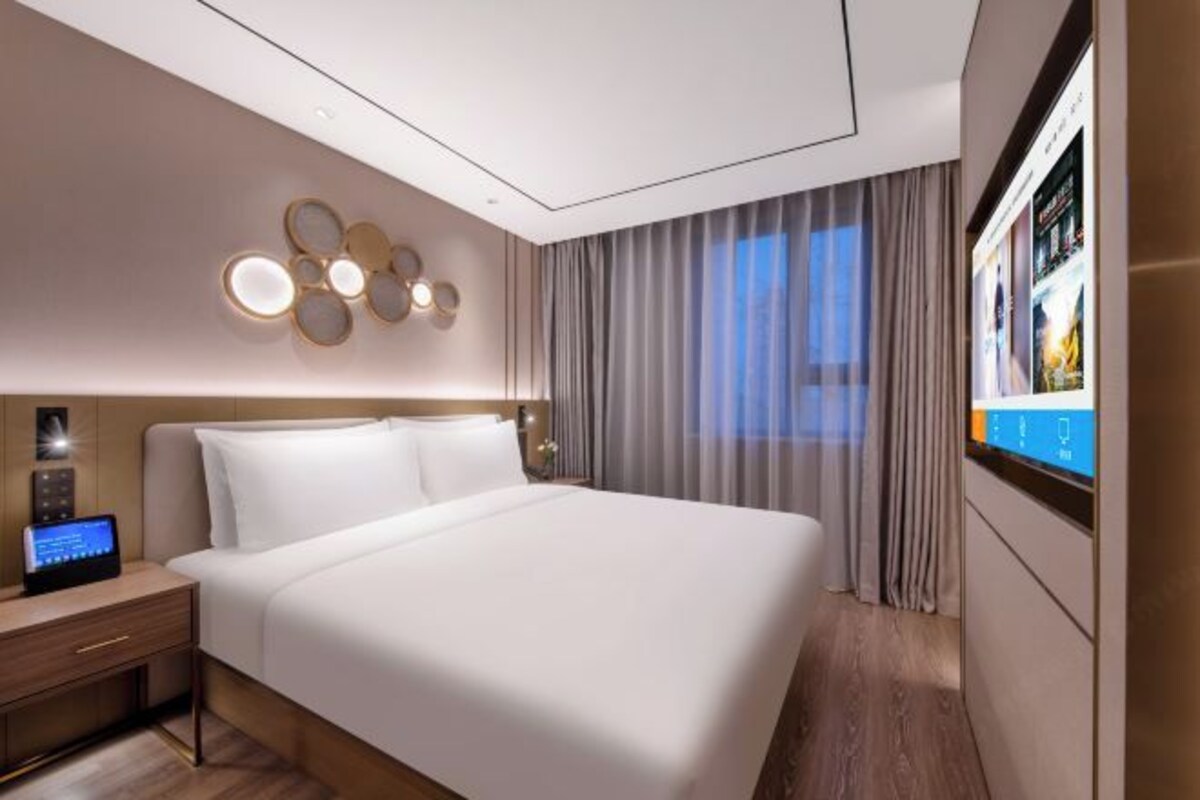 Ein modernes Hotelzimmer mit einem großen Bett, einem Nachttisch mit Bedienfeld, einer Wandleuchte und einem großen Fenster mit Vorhängen.