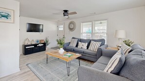A modern living room with a grey sofa, a wooden coffee table, a black entertainment unit, a ceiling fan, and a large window with blinds. - Charming duplex with balcony,  W/D, central AC & steps to beach (Murrells Inlet)