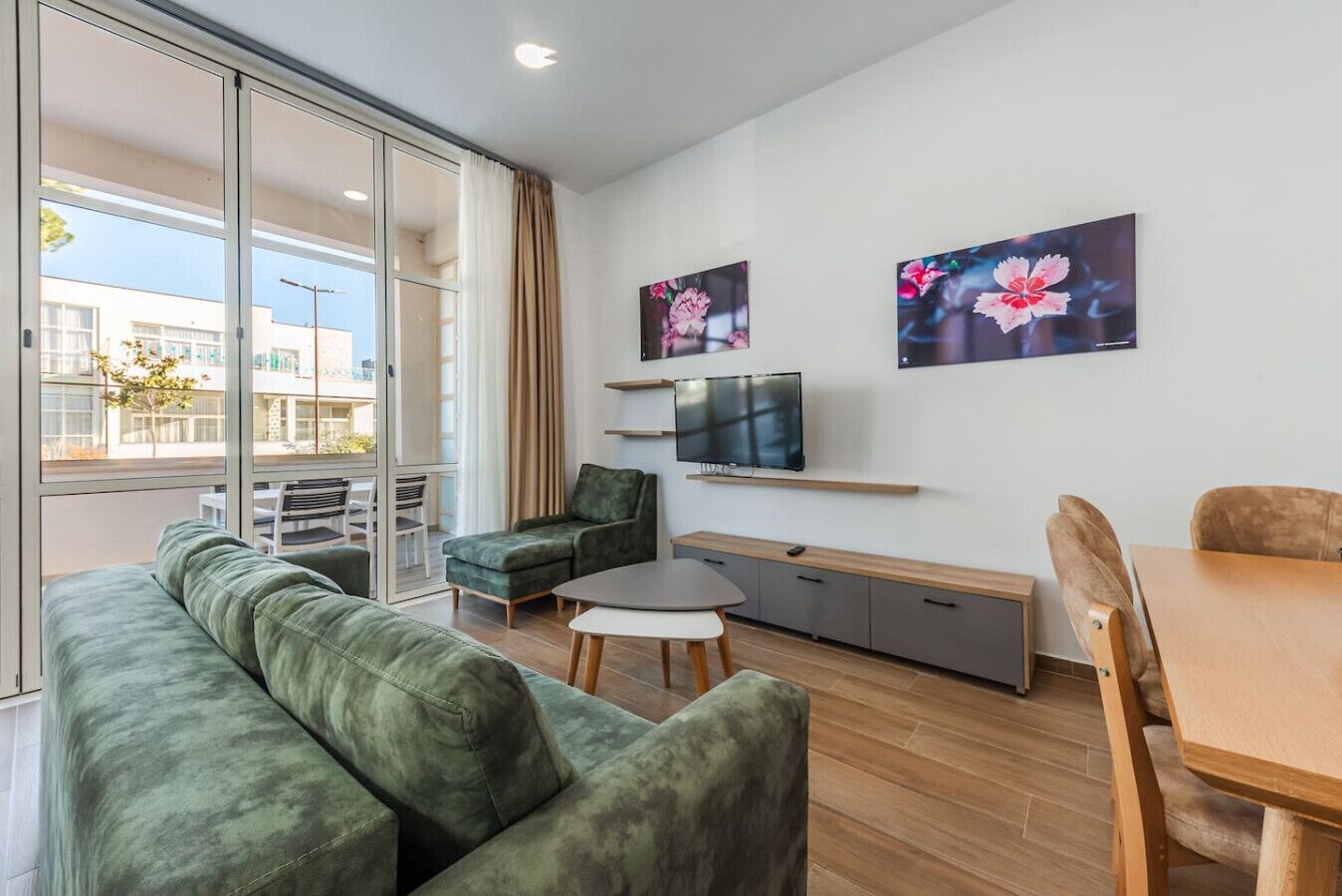 A modern living room with a green sofa, a small table, a TV on a shelf, and a dining area with wooden chairs.