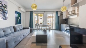 Un salon moderne avec un canapé gris, une table à manger avec des chaises, une cuisine équipée d’une cuisinière et d’un four, et deux lampes suspendues.