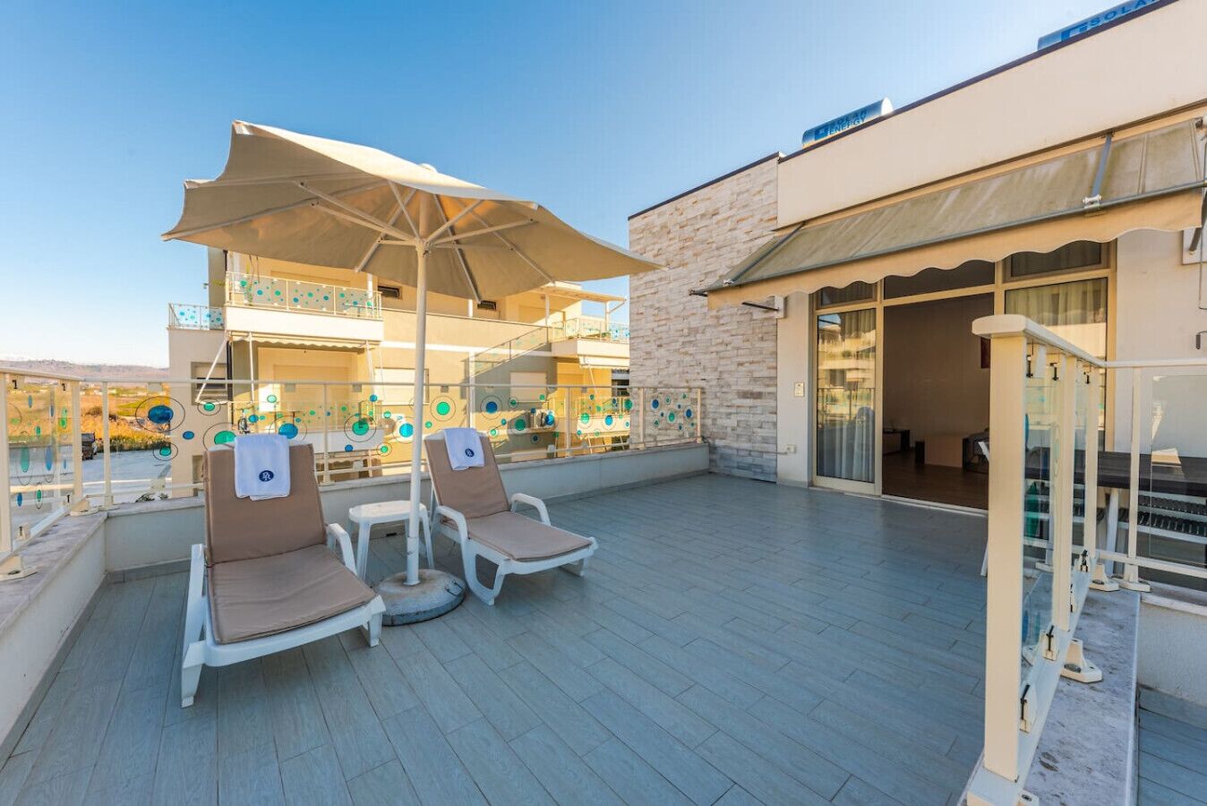 A rooftop terrace with lounge chairs and a large umbrella, overlooking a pool area with a stone wall and glass doors.