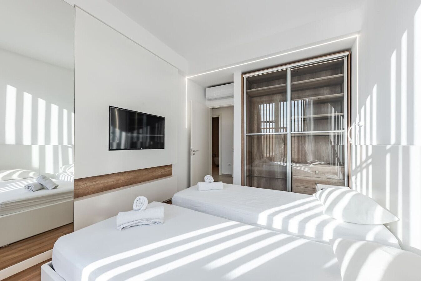 Habitación de hotel con dos camas, televisión, un espejo grande y puerta corrediza de vidrio que da a un balcón.