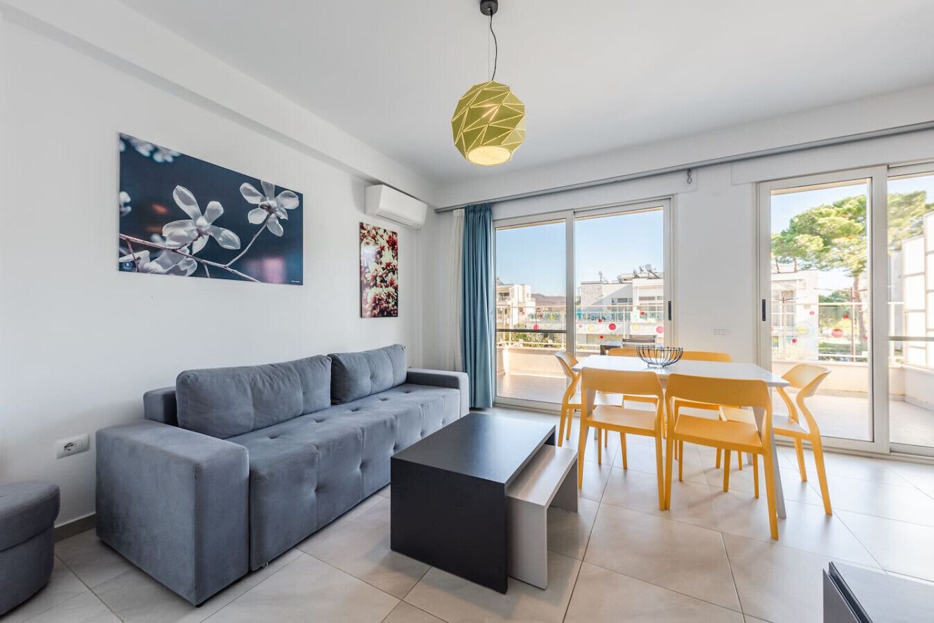 A modern living room with a grey sofa, a black coffee table, and a dining area with yellow chairs. There are large windows offering a view of the outside.