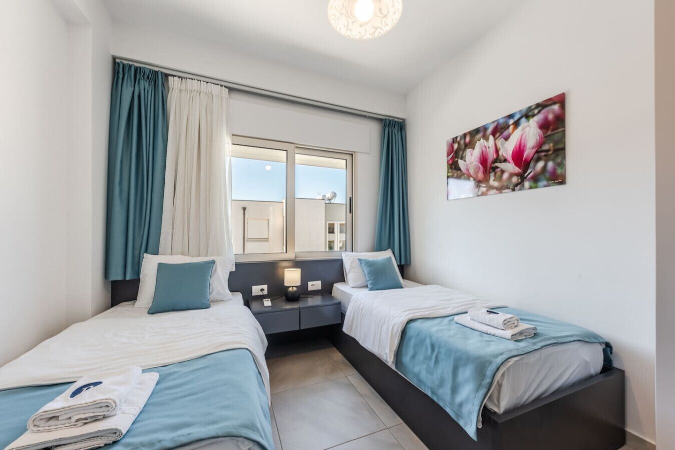 Ein Doppelzimmer mit zwei Betten, einem Fenster mit Vorhängen, einem Nachttisch mit Lampe und einem Blumenbild an der Wand.