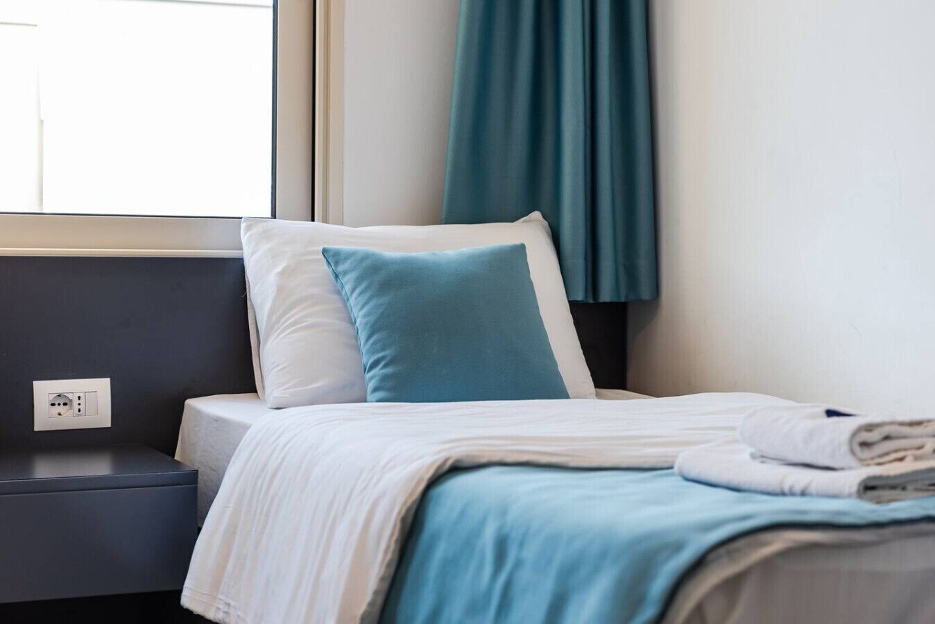 A neatly made bed with white and blue bedding, a dark headboard, and a window with curtains.