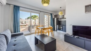 A modern living room with a sofa, dining table, and chairs, a kitchen area with a TV and appliances, and large windows with curtains. - Tranquil Apartment with pool access - Manjola by PikHost (Gjiri i Lalzit)