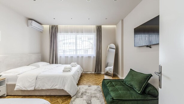 A modern hotel room with a large bed, a green sofa, a TV mounted on the wall, and a large window with curtains. - 1 Bedroom Loft - Serenitas 1 by PikHost (Tiranë)