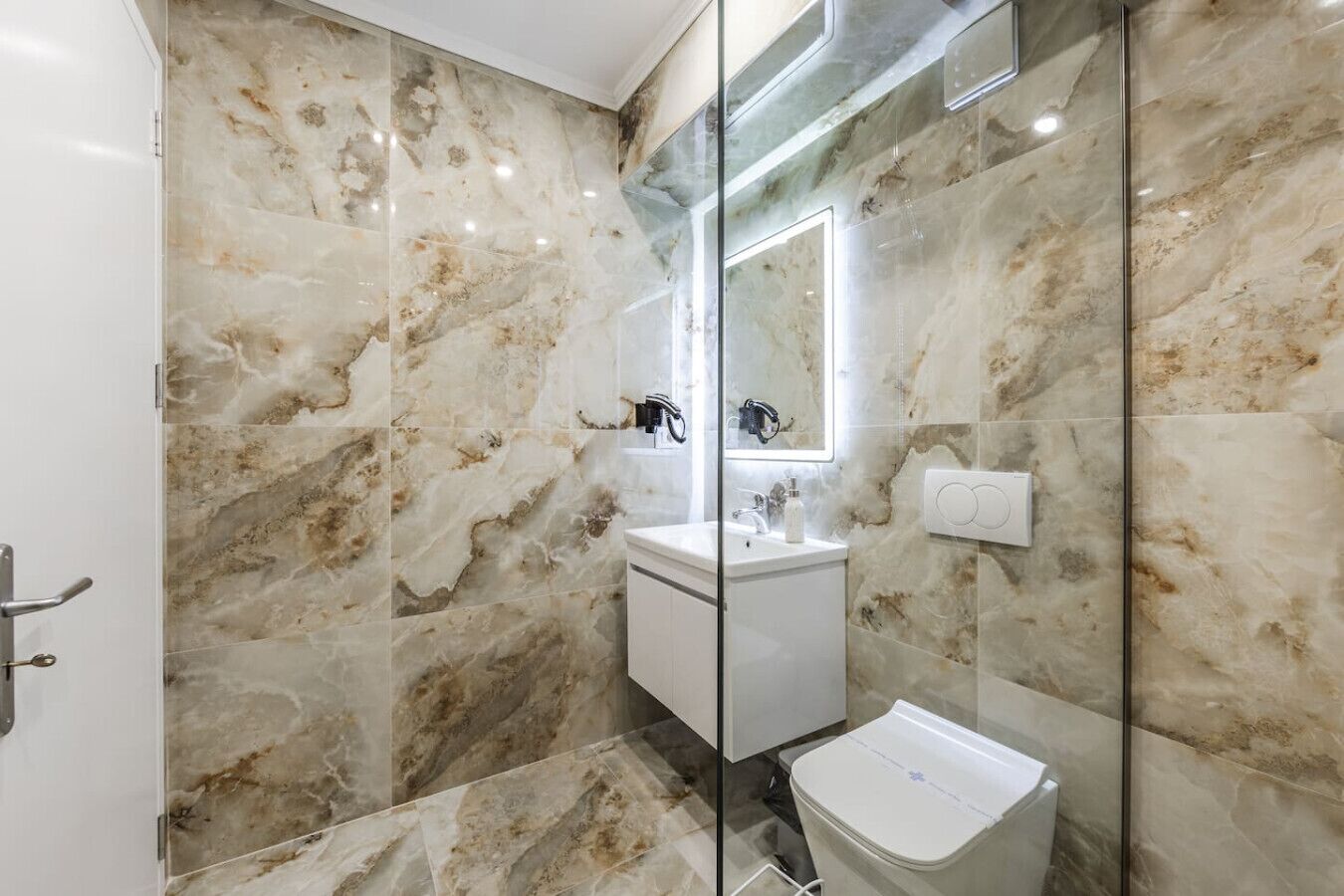 A bathroom with marble walls, a glass shower enclosure, a white sink, and a toilet.