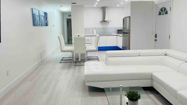A modern living room with a white sectional sofa, a glass coffee table, and a dining area with white chairs. - Beautiful apartment two mins away from the beach! (Fort Myers Beach)