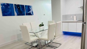 A modern dining area with a glass table and white chairs, a kitchen with white cabinets, and blue abstract art on the wall.