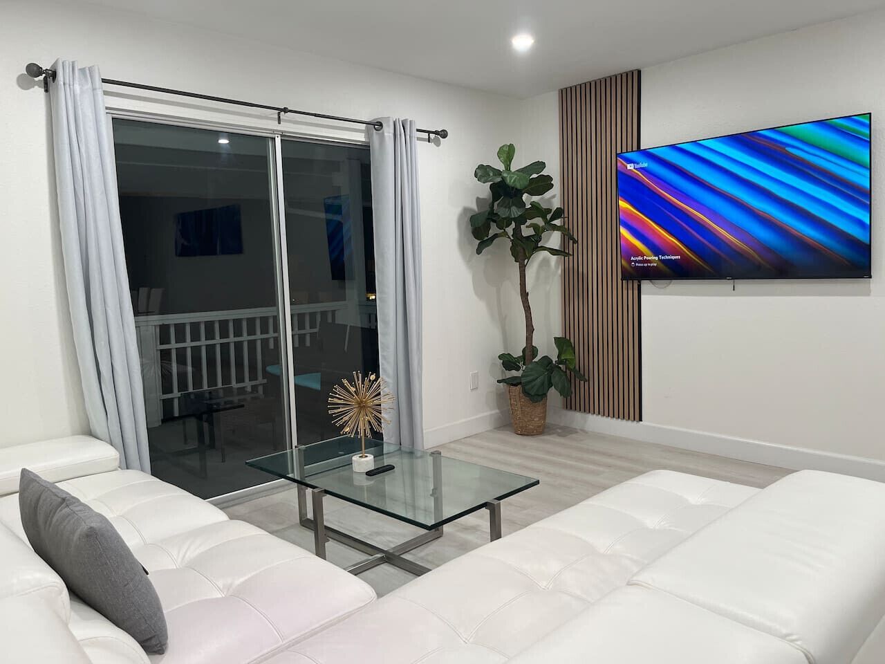 A modern living room with a glass coffee table, white sofas, a large flat-screen TV, a sliding glass door, and a potted plant.