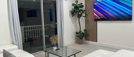 A modern living room with a glass coffee table, white sofas, a large flat-screen TV, a sliding glass door, and a potted plant.