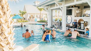 A pool area with people swimming and relaxing, a bar with various items on display, and a building with a distinctive roof and columns in the background. - Luxurious four bedroom beach home with patio, balconies, firepit & game room (Port Aransas)