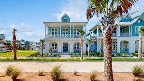A large, two-story beachfront house with a balcony, palm trees, and a clear sky. - Luxurious four bedroom beach home with patio, balconies, firepit & game room (Port Aransas)