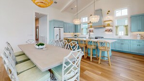 A spacious kitchen with light blue cabinets, a central island with bar stools, and a dining area with white chairs and a table. - Luxurious four bedroom beach home with patio, balconies, firepit & game room (Port Aransas)