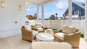 A covered outdoor seating area with wicker furniture and a glass-top table, overlooking a residential neighborhood. - Luxurious four bedroom beach home with patio, balconies, firepit & game room (Port Aransas)