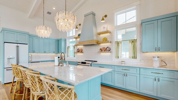 A modern kitchen with light blue cabinets, a large island with bar stools, a stainless steel refrigerator, and a pendant light fixture. - Luxurious four bedroom beach home with patio, balconies, firepit & game room (Port Aransas)