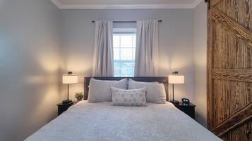 Un dormitorio con una cama grande, dos mesitas de noche con lámparas, una ventana con cortinas y un panel de madera en la pared derecha.