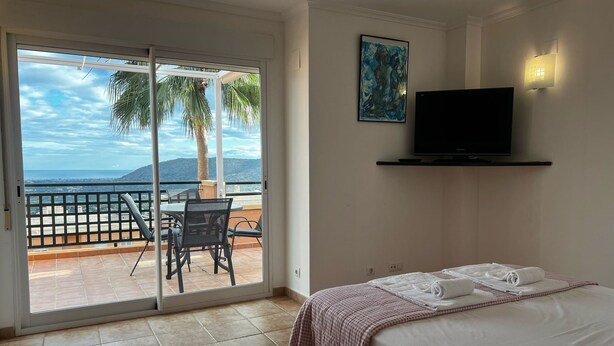 A hotel room with a bed, a TV, a painting, and a balcony with a view of the sea and palm trees. - Jacaranda in Pedreguer (Pedreguer)