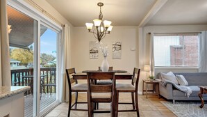 A dining area with a chandelier, a round table, and four chairs. There is a sofa and a window with blinds in the adjacent living area. - Brighton House - Walk to Metro (Seattle)