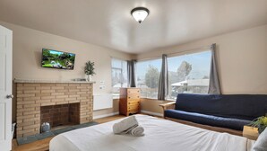 A hotel room with a brick fireplace, a bed with white bedding, a blue sofa, a television on the wall, and a window with curtains. - Brighton House - Walk to Metro (Seattle)