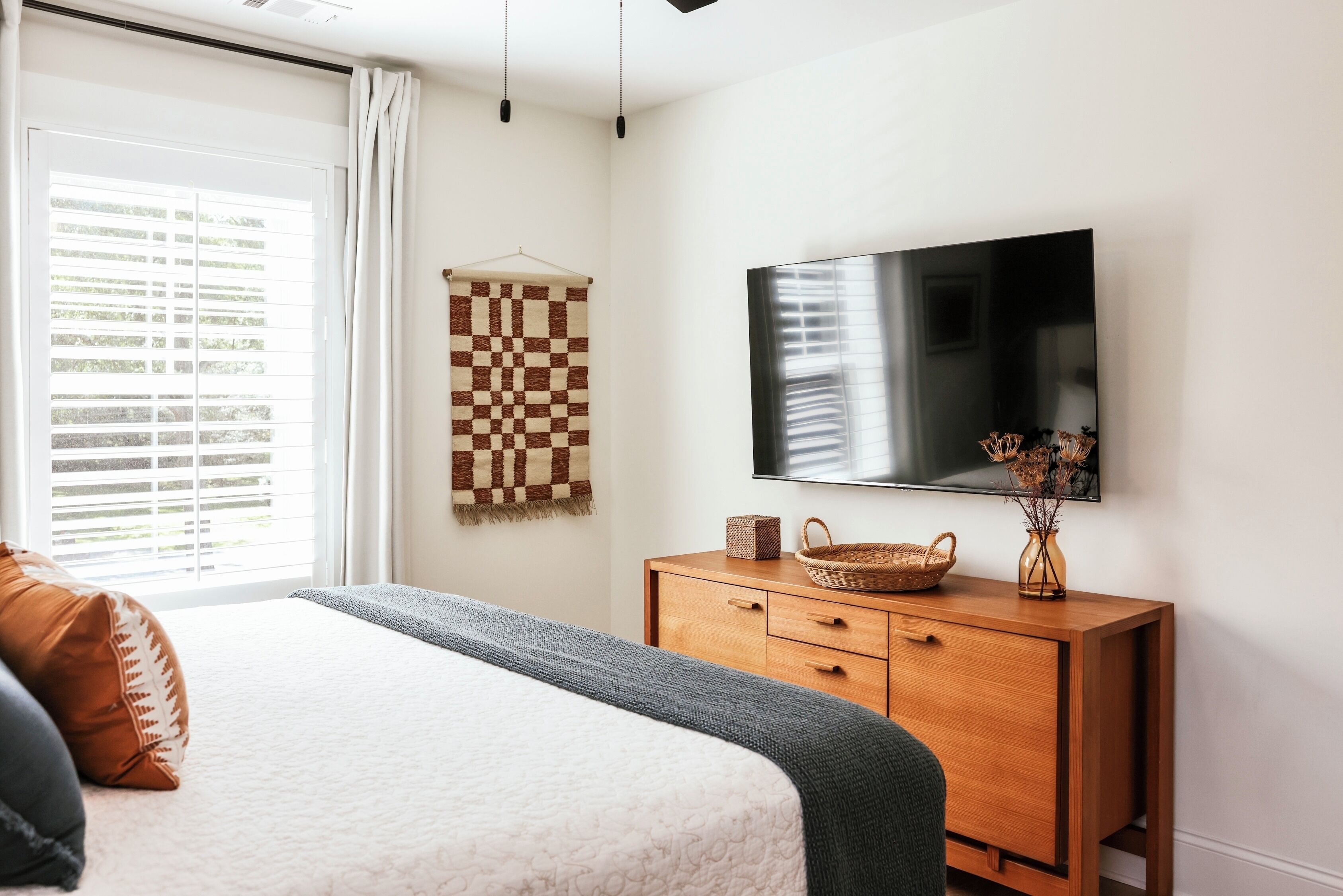 A bedroom with a bed, a wooden dresser, a television, and a window with blinds.