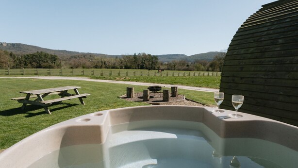 A hot tub with a picnic table and two wine glasses, set against a backdrop of a grassy field and distant hills.