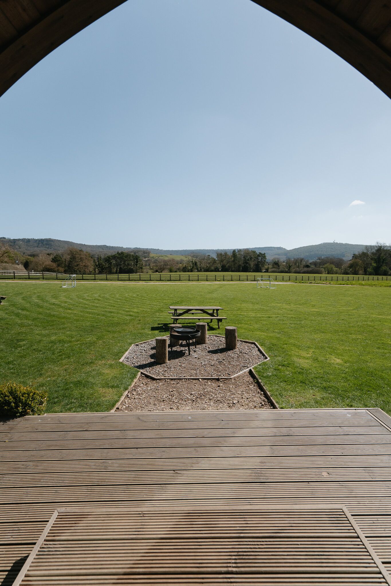 A wooden deck with picnic tables and a barbecue grill, overlooking a grassy field with a fence and trees in the distance.