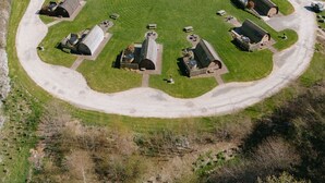 Vue aérienne d’une propriété rurale avec un grand champ verdoyant, une route sinueuse et plusieurs petits bâtiments aux toits distinctifs.