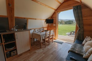 A cozy, wooden interior with a dining area, kitchen, and a sofa by a large window overlooking a grassy landscape.