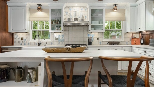 Een keuken met witte kastjes, een centraal eiland met twee houten stoelen en een raam met uitzicht op groen.
