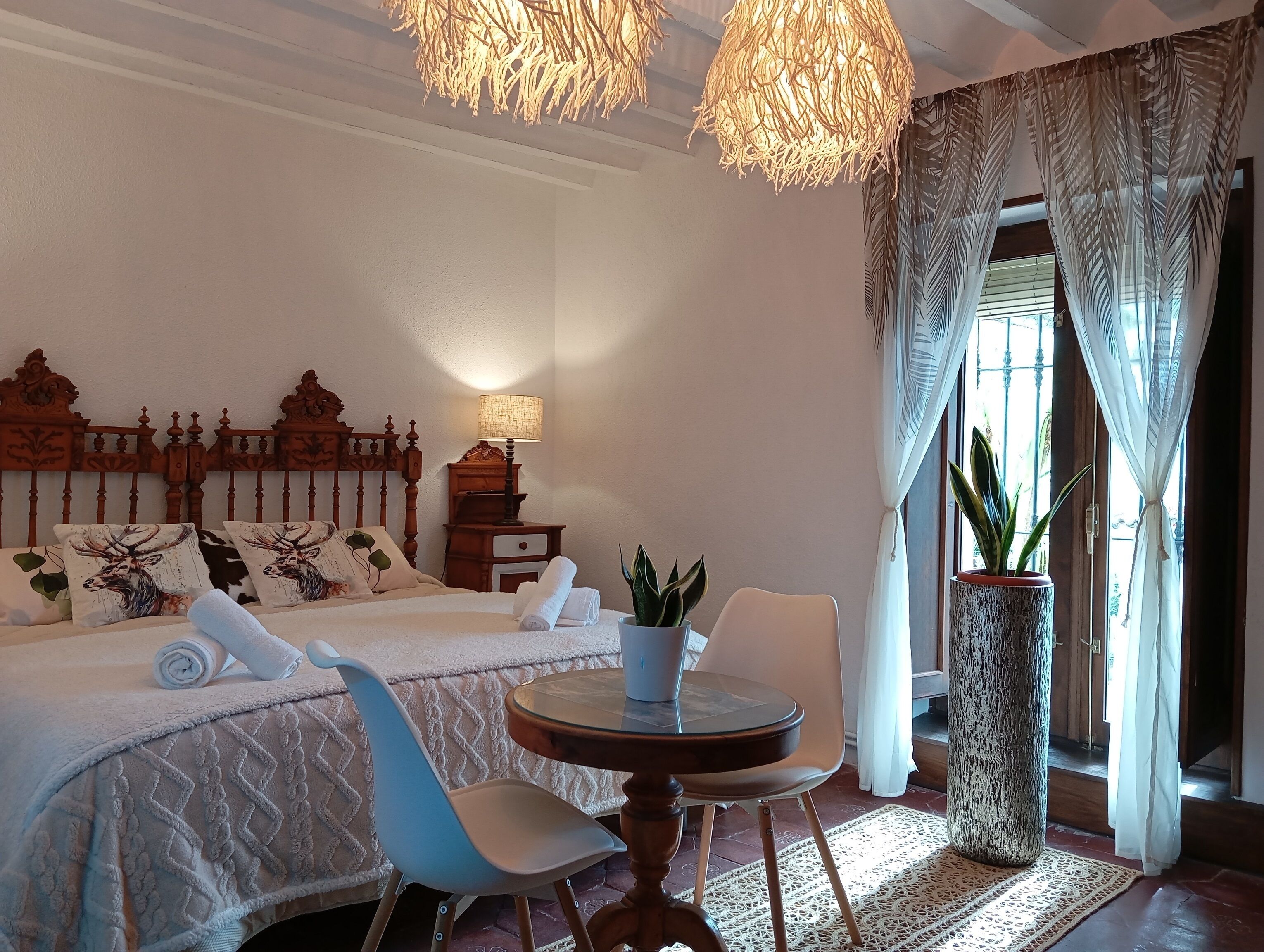 A bedroom with a large bed, a bedside table, a lamp, a chair, a round table, and a potted plant.