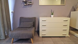 Ein Schlafzimmer mit grauem Sofa, einem weißen Schminktisch, einem Spiegel und einem Wandregal mit Pflanzen.